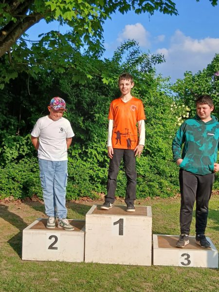 Three children stand on a podium in an outdoor setting. The child on the first place podium (center) wears an orange shirt and black pants, smiling. The second place child (left) is in a white shirt and blue pants, with a colorful cap. The third place child (right) wears a green and black patterned sweatshirt and black pants. The background features lush greenery and a partly cloudy sky.