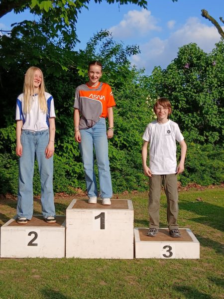 Three children stand on a podium in an outdoor setting. The child on the left, wearing a white and blue shirt, is on the second place platform. The child in the middle, wearing an orange and gray shirt, stands on the first place platform. The child on the right, dressed in a white shirt, is on the third place platform. The background features green foliage and a blue sky with some clouds.