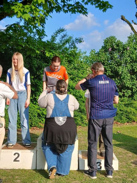 A group of people is standing on a podium in an outdoor setting. Two individuals, a girl with long blonde hair and another with a short hairstyle, are positioned on the first and second steps of the podium, respectively. A person wearing a black shirt and denim overalls is on the third step, facing away from the camera. Another individual, dressed in a striped shirt, is interacting with someone off to the side. The background features lush green foliage and a clear blue sky.