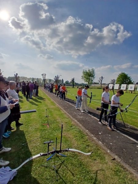 A sunny outdoor archery range is depicted, with a long path lined by participants preparing to shoot. Archers are seen in various positions, some holding bows and others adjusting their equipment. In the background, several targets are set up on stands. The area is surrounded by neatly trimmed grass and trees, with a few clouds in the blue sky. Spectators are gathered along the sidelines, observing the event.