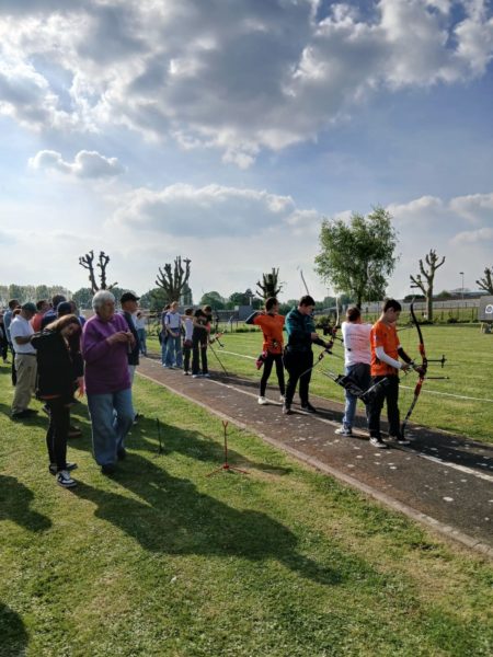 A group of archers is walking along a path in a grassy area, preparing for an archery event. Some participants are wearing bright orange shirts, while others are in darker clothing. They are carrying bows and arrows, and a few spectators are gathered nearby, observing the activity. The sky is partly cloudy, and there are trimmed trees in the background, adding to the outdoor setting.