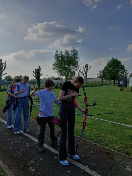 A group of four young people is lined up on a grassy archery range, each holding a bow. They appear focused on their equipment, with some adjusting their bows. The background features a clear blue sky with scattered clouds and a few trees. In the distance, an archery target is visible. The scene conveys a sense of practice and concentration in an outdoor setting.