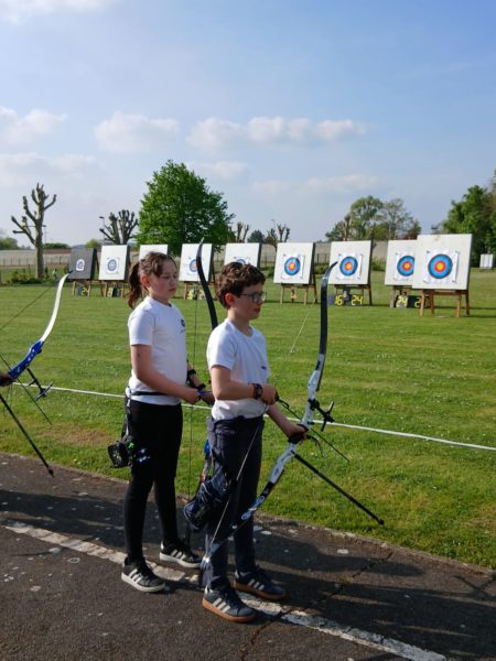 Two young archers stand on a grassy field, each holding a bow. They are dressed in matching white shirts and dark pants. In the background, several archery targets are set up, displaying colorful bullseyes. The sky is clear with a few clouds, and a tree is visible nearby. The atmosphere appears calm and focused, typical of an archery practice session.