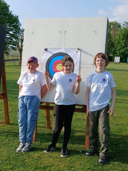 Three children stand in front of an archery target on a grassy field. The target features concentric circles in blue, red, and yellow. The child on the left wears a cap and a white t-shirt, smiling. The girl in the center, also in a white t-shirt, makes a peace sign with her fingers. The child on the right wears glasses and a white t-shirt, standing casually. The background includes trees and a clear sky.