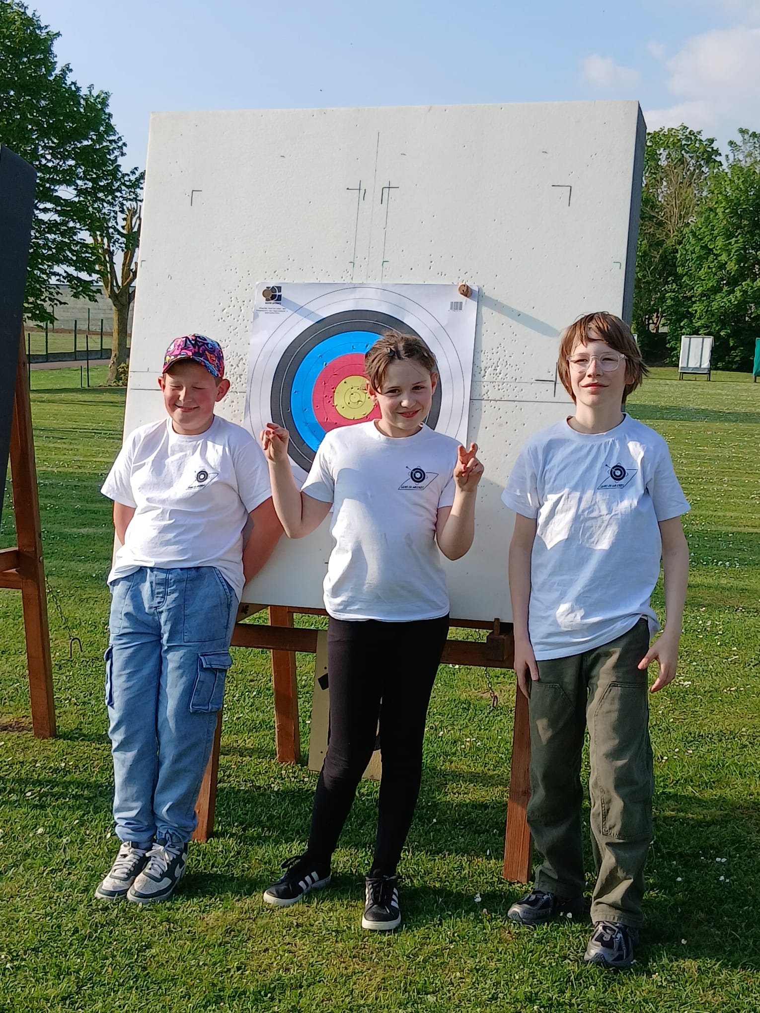 Three children stand in front of an archery target on a grassy field. The target features concentric circles in blue, red, and yellow. The child on the left wears a cap and a white t-shirt, smiling. The girl in the center, also in a white t-shirt, makes a peace sign with her fingers. The child on the right wears glasses and a white t-shirt, standing casually. The background includes trees and a clear sky.