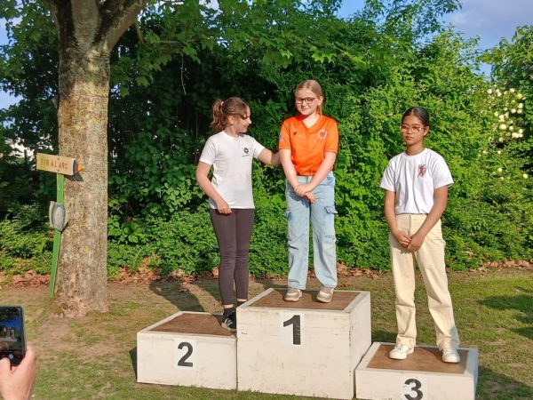 Three young girls stand on a podium, celebrating their achievements. The girl in the center, wearing an orange shirt and light blue pants, occupies the first place position. To her left, a girl in a white shirt and black pants stands in second place, while another girl in a white shirt and beige pants is in third place on the right. In the background, there is a tree and greenery, along with a sign that reads "TIR À L'ARC." The setting appears to be outdoors, likely at a sports event.