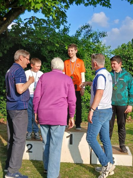A group of people is gathered around a podium in an outdoor setting. A young boy wearing an orange shirt stands on the first place podium, smiling and holding a medal. Two other boys, one in a white shirt and the other in a green patterned shirt, stand on the second and third place podiums, respectively. An older woman in a purple sweater is facing the boy on the first podium, while two men, one in a blue striped shirt and the other in a white shirt with blue accents, are engaged in conversation with the group. The background features green foliage and a clear blue sky.