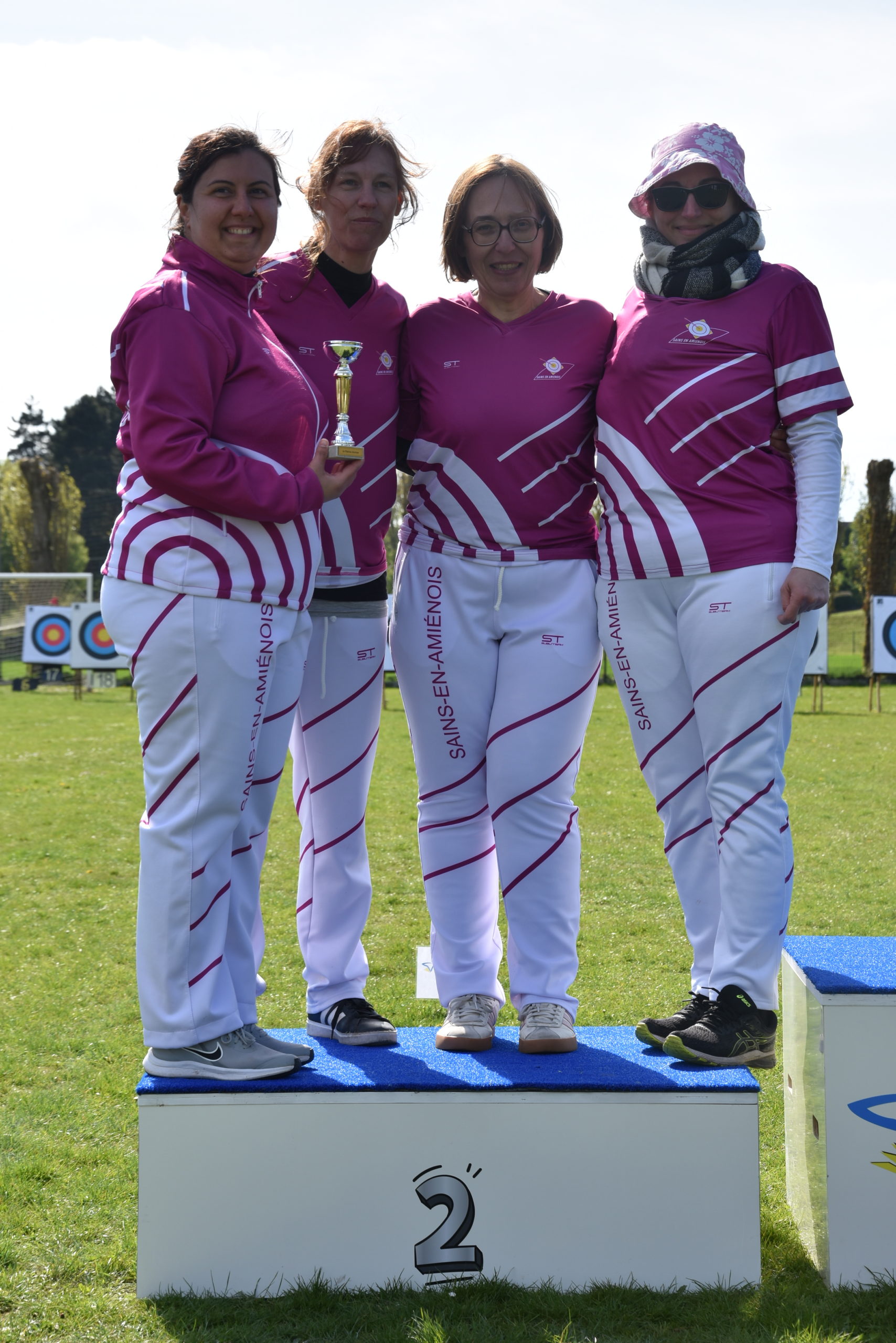 Four women stand on a podium, celebrating a second-place finish in an archery competition. They are wearing matching pink and white athletic outfits, with the words "SANS-EN-AMÉNOIS" printed on their pants. One woman holds a small trophy, while the others smile and pose together. The background features a grassy field with archery targets visible in the distance, under a clear sky.