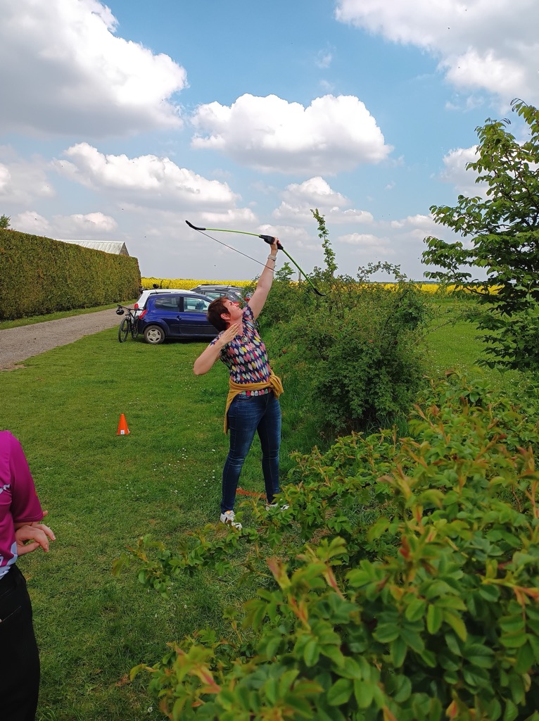 A person is standing outdoors, aiming a bow with an arrow drawn back. They are wearing a colorful, patterned shirt and blue jeans, with a yellow belt around their waist. In the background, there is a blue car parked on a gravel path, and a grassy area with some bushes and trees. The sky is partly cloudy, and there is a small orange cone visible on the ground nearby.