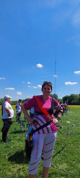 A woman stands confidently on a grassy field, wearing a bright pink and white archery outfit with the text "SAINS-EN-AMIENOIS" on her pants. She has a red sash across her chest and is equipped with an archery bow. In the background, other archers are visible, and the sky is clear with a few clouds. The setting appears to be an outdoor archery event.