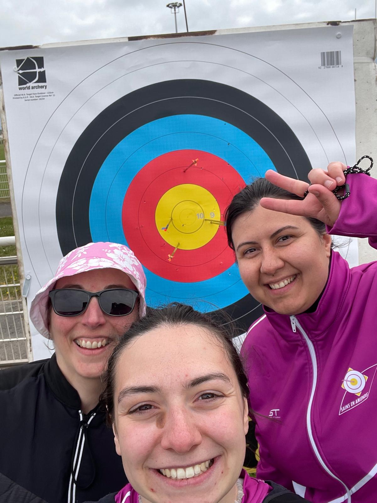 Three individuals are smiling in front of an archery target. The target features concentric circles in blue, black, red, and yellow, with arrows embedded in the red and yellow areas. One person is wearing sunglasses and a pink hat, while another is making a peace sign. The third person is dressed in a purple jacket. The background shows a cloudy sky and a sports facility.