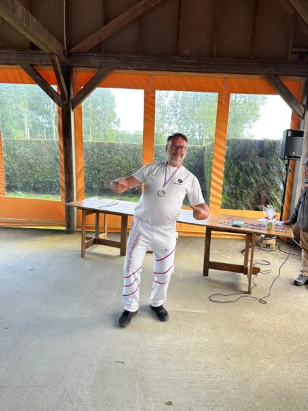 A man stands in a covered outdoor area, smiling and posing with a playful gesture. He wears a white t-shirt and white pants with pink stripes, along with a medal around his neck. In the background, there are large windows with orange curtains, revealing greenery outside. A wooden table is set up nearby, and another person can be seen in the background, partially obscured. The floor is concrete, and the overall atmosphere appears casual and celebratory.