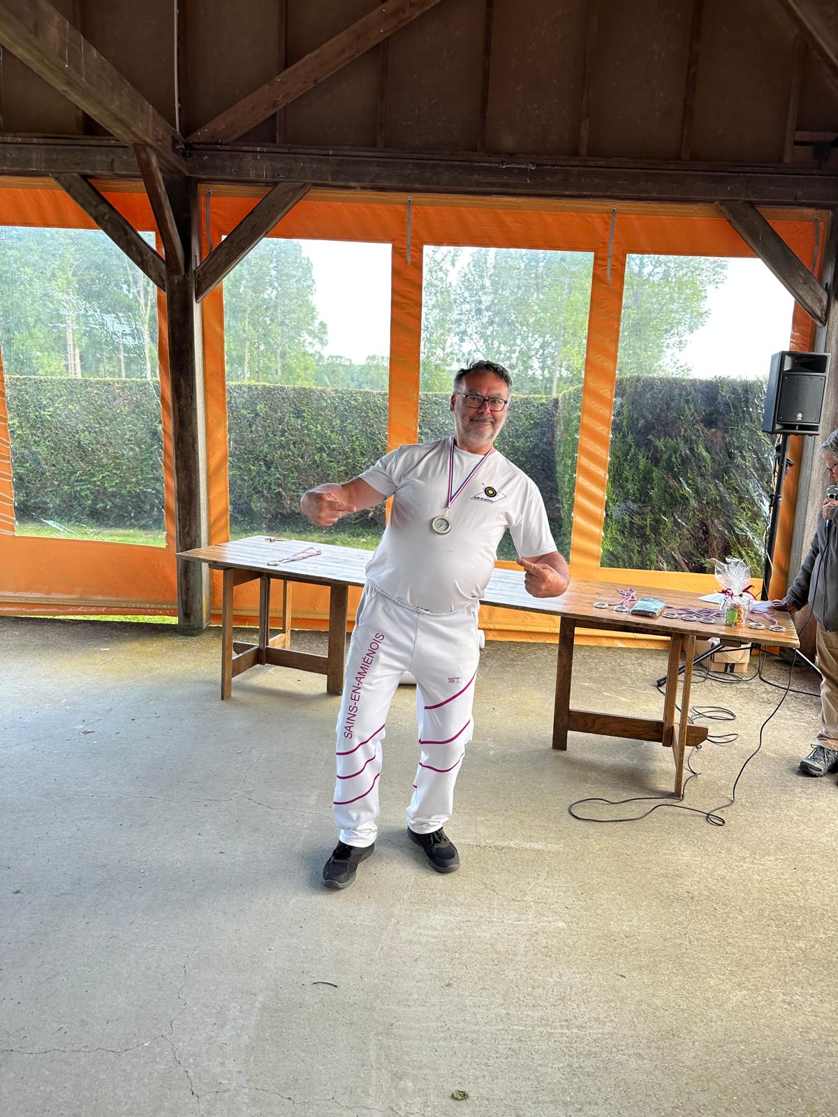 A man stands in a covered outdoor area, smiling and posing with a playful gesture. He wears a white t-shirt and white pants with pink stripes, along with a medal around his neck. In the background, there are large windows with orange curtains, revealing greenery outside. A wooden table is set up nearby, and another person can be seen in the background, partially obscured. The floor is concrete, and the overall atmosphere appears casual and celebratory.