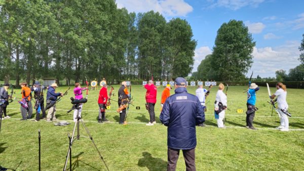 A group of archers is lined up on a grassy field, preparing to shoot at targets in the distance. They are dressed in a variety of colorful outfits, including bright orange, red, and blue. Some archers are holding their bows in shooting position, while others are adjusting their gear. In the foreground, an official wearing a navy jacket with "ARBITRE" written on the back is overseeing the event. The background features several archery targets set up on stands, surrounded by trees under a partly cloudy sky.