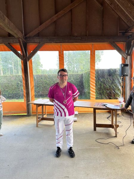 A person stands in a spacious indoor area with orange curtains and wooden beams. They are wearing a bright pink shirt with white stripes and white pants featuring a design. A medal hangs around their neck. In the background, there are tables with items on them, and a speaker is visible. The setting appears to be a casual event or gathering.