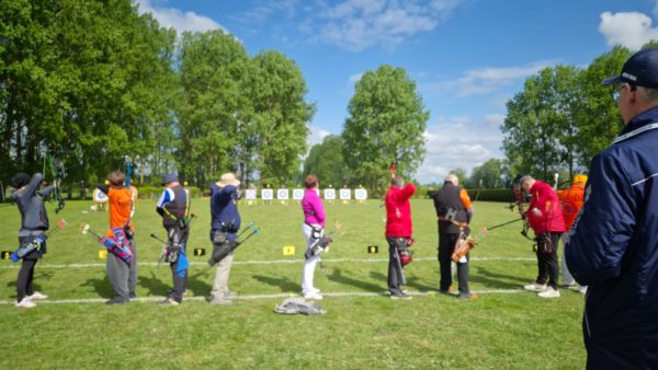 A group of archers is lined up on a grassy field, preparing to shoot at targets in the distance. Each archer is holding a bow, with arrows secured in quivers attached to their sides. The archers are dressed in a variety of colorful clothing, including orange, red, and purple. In the background, several archery targets are visible, marked with concentric circles. The sky is partly cloudy, and there are tall trees lining the area, creating a serene outdoor setting.