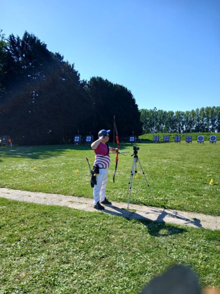 A person stands on a grassy field, holding a bow in one hand and a camera on a tripod in the other. They are dressed in a sleeveless pink shirt and white pants with red stripes. The background features several archery targets arranged in a row, with trees lining the area under a clear blue sky. The ground is marked with small cones, and the scene is well-lit, suggesting a sunny day.