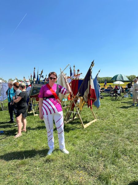 A woman stands in a grassy field under a clear blue sky, wearing a vibrant pink and white outfit with a sash. She is posing with one hand raised, surrounded by colorful banners and flags displayed on wooden stands. In the background, other people are visible, some seated and others standing, enjoying the event. The scene conveys a festive atmosphere.