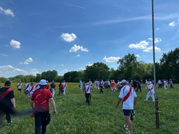 A large group of people is walking across a grassy field under a bright blue sky with scattered clouds. Many participants are wearing white shirts with red sashes, while others are in red shirts. The scene is lively, with individuals moving in various directions, and a few trees are visible in the background. A tall pole stands on the right side of the image, suggesting an organized event or gathering.
