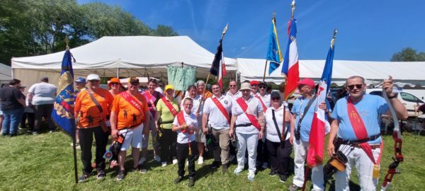 A group of individuals stands together outdoors, smiling and posing for the camera. They are dressed in a variety of colorful outfits, including orange and white shirts, and some wear sashes. Several members hold flags, showcasing different colors and designs, while one person holds a bow. In the background, a large white tent is visible, along with other people and greenery. The sky is clear and blue, indicating a sunny day.