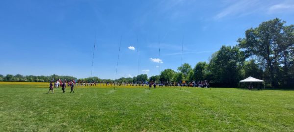 A wide grassy field under a clear blue sky is filled with groups of people standing in formation. They are holding various flags, with some individuals walking in front of the formations. In the background, there are trees lining the edge of the field, and a white tent is set up to the right, suggesting a gathering or event. The atmosphere appears festive and organized.