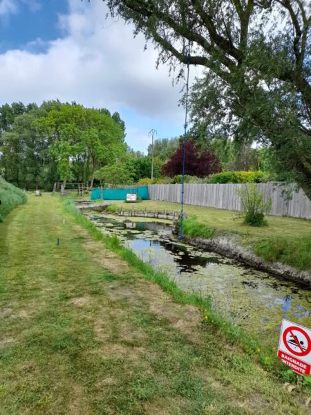 A grassy path runs alongside a narrow, calm waterway, bordered by trees and shrubs. On the left, a wooden structure extends into the water, while a sign indicating "No Swimming" is prominently displayed on the right. A green tarp is visible in the background, and the sky is partly cloudy. The overall scene is tranquil and natural, with a mix of greenery and water.