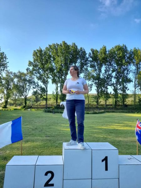 A woman stands on a podium, specifically on the first place section, which is marked with the number "1." She is wearing a white t-shirt and blue jeans, holding a medal in her hands. The background features a grassy area with several trees and a clear blue sky. To her left, there is a blue and white flag, and to her right, a flag with a design that includes the colors of the Union Jack. The second place section of the podium is marked with the number "2.