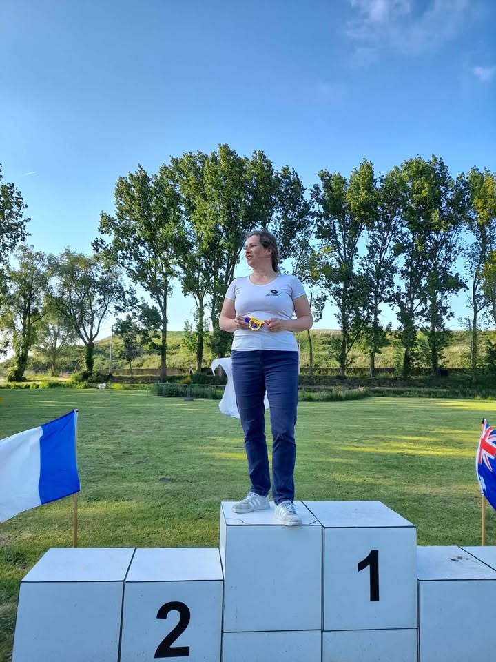 A woman stands on a podium, specifically on the first place section, which is marked with the number "1." She is wearing a white t-shirt and blue jeans, holding a medal in her hands. The background features a grassy area with several trees and a clear blue sky. To her left, there is a blue and white flag, and to her right, a flag with a design that includes the colors of the Union Jack. The second place section of the podium is marked with the number "2.