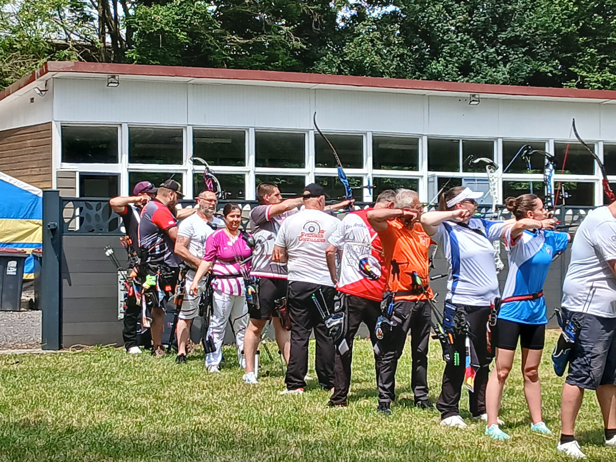 A group of archers is lined up on a grassy field, preparing to shoot. They are wearing various colored shirts, including pink, orange, and blue, and each has a quiver attached to their waist. The archers are in a stance with their bows drawn back, focused on their targets. In the background, a building with large windows and a colorful tent is visible, surrounded by trees. The scene is bright and sunny, indicating a pleasant day for archery practice.