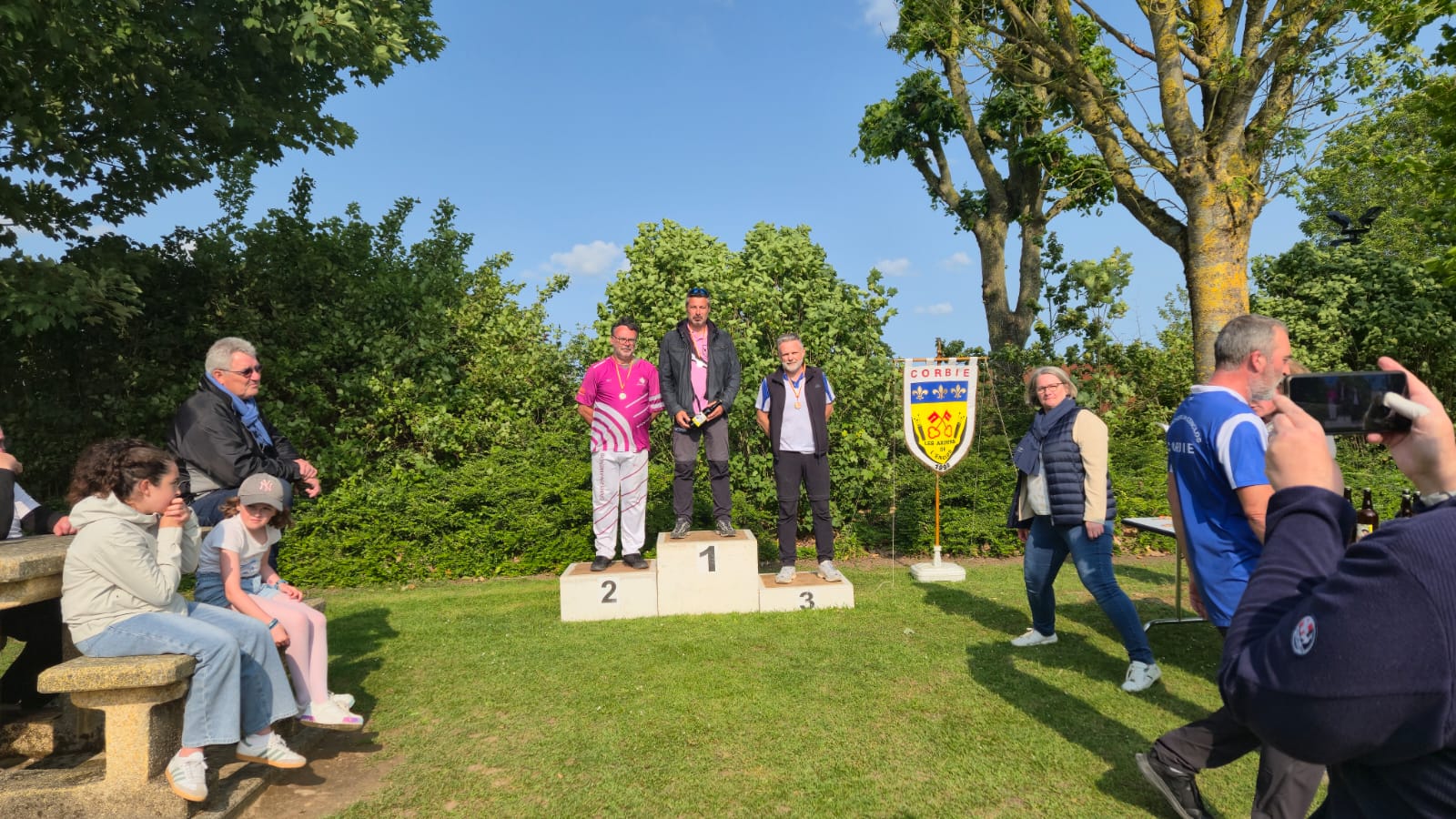 Three individuals stand on a podium, celebrating their achievements in a competition. The person in the center, wearing a black jacket, holds a trophy and is flanked by two others: one in a pink shirt and the other in a blue and white shirt. The podium is marked with numbers 1, 2, and 3, indicating their placements. In the background, there are trees and a banner displaying a coat of arms. A small group of spectators, including children and adults, is seated on a stone bench nearby, while others are walking around, some taking photos.
