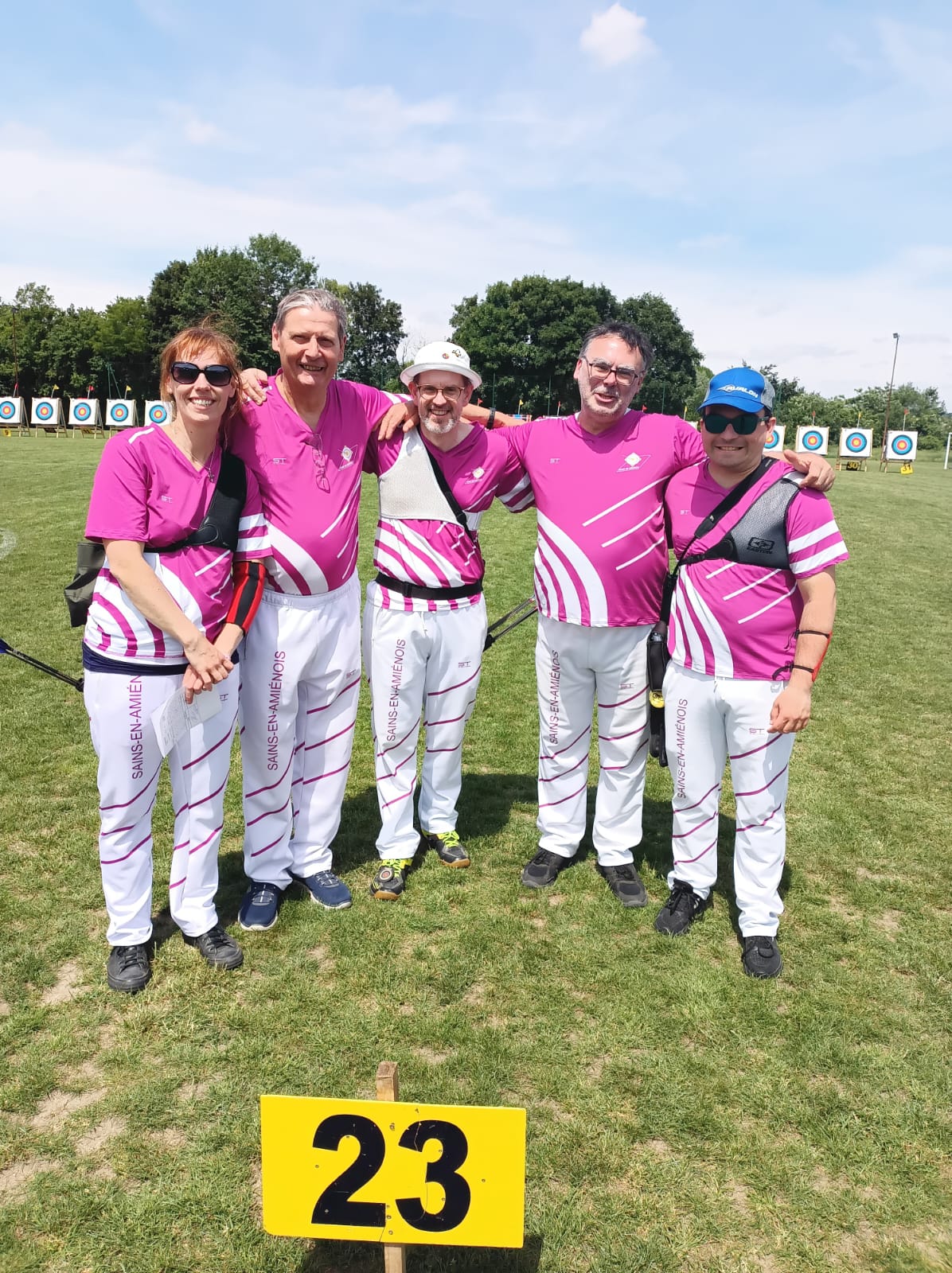 A group of five archers stands together on a grassy field, smiling and posing for the photo. They are wearing matching pink and white uniforms with a design that includes diagonal stripes. The background features several archery targets set up in a row. A yellow sign with the number "23" is placed in front of them on the ground. The sky is clear with a few clouds, indicating a sunny day.