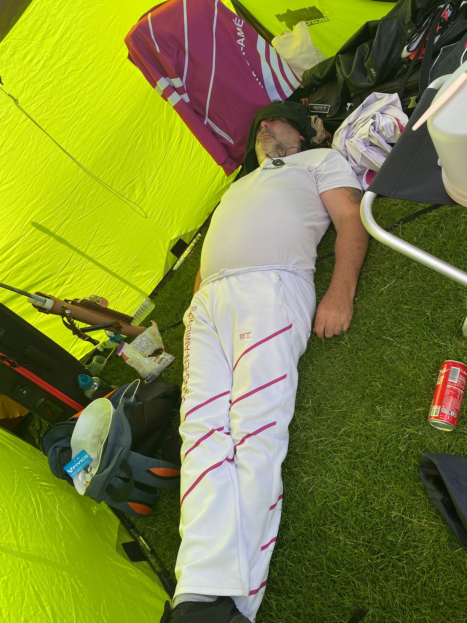 A person is lying on the grass inside a bright yellow tent. They are wearing a white shirt and white pants with pink stripes. The individual has a black cloth over their eyes, suggesting they might be resting or sleeping. Surrounding them are various items, including a backpack, clothing, and a can of soda. The tent's interior is illuminated by natural light, creating a vibrant atmosphere.